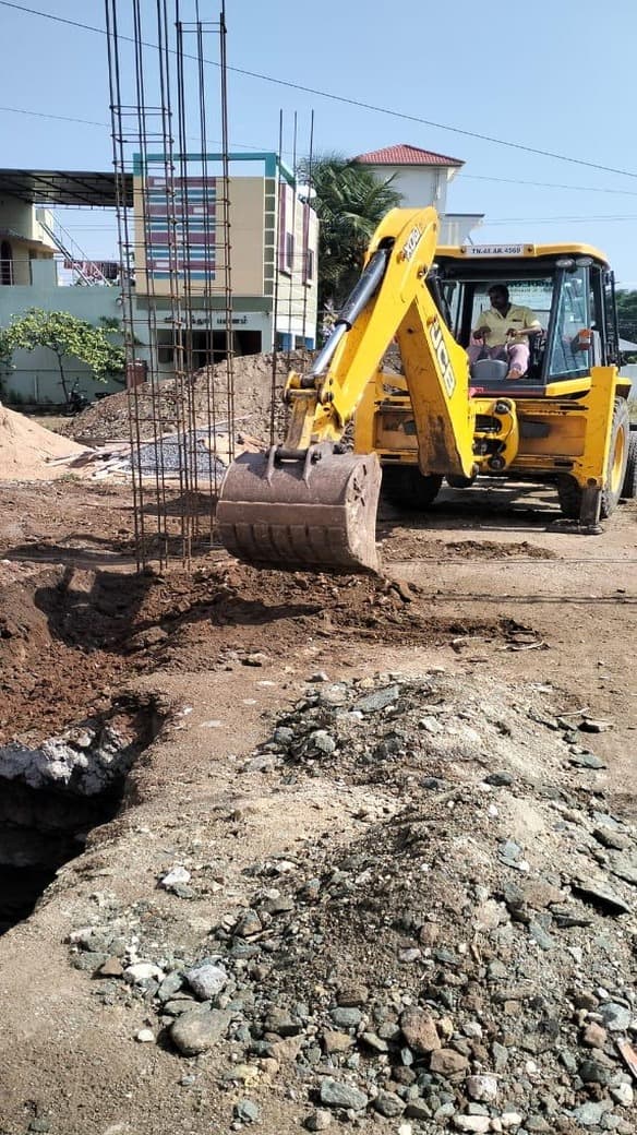 A backhoe excavator cutting foundation trenches beside standing column cages on a residential site.