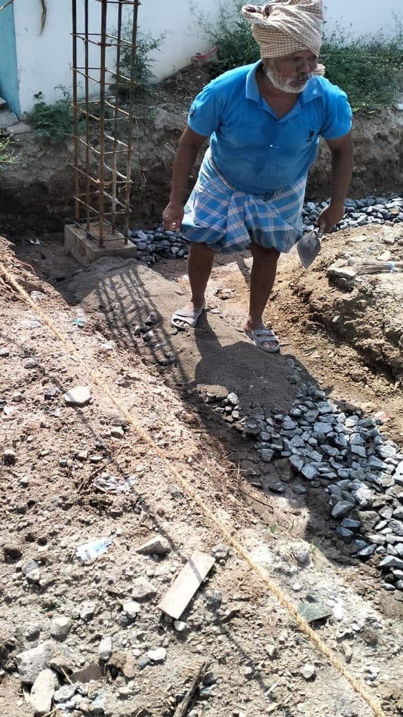 A mason with a trowel working beside a column reinforcement cage and a heap of graded aggregate.